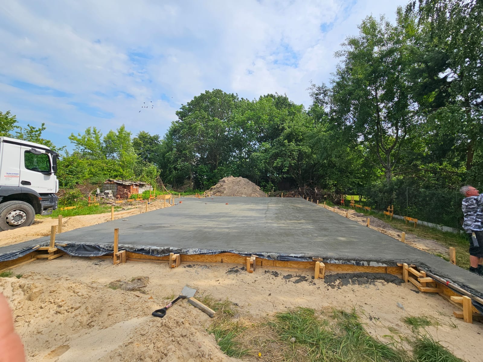 Wylana płyta fundamentowa z drewnianym szalunkiem na piaszczystym terenie, widoczny biały samochód ciężarowy i sterta ziemi w tle. Słoneczny dzień.