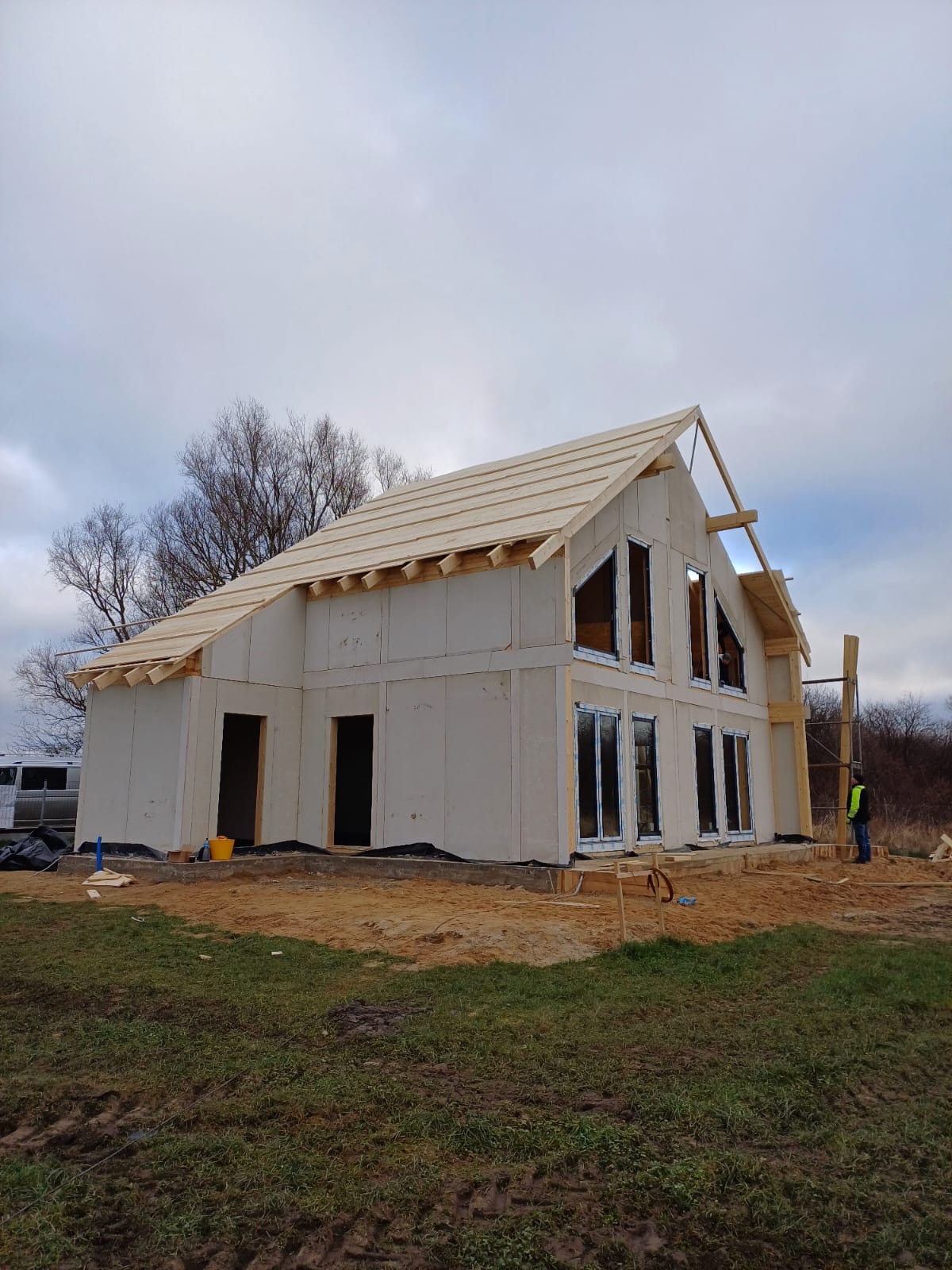 Budowa domu szkieletowego w trakcie: ściany z płyt OSB, drewniana konstrukcja dachu, wstawione okna. Na zewnątrz piasek, trawa i pracownik w kamizelce.