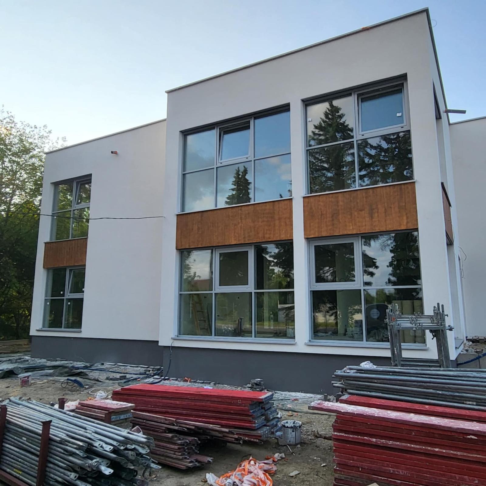 Nowoczesny, dwukondygnacyjny budynek z dużymi oknami i drewnianymi panelami elewacyjnymi. Na pierwszym planie leżą materiały budowlane i rusztowania.