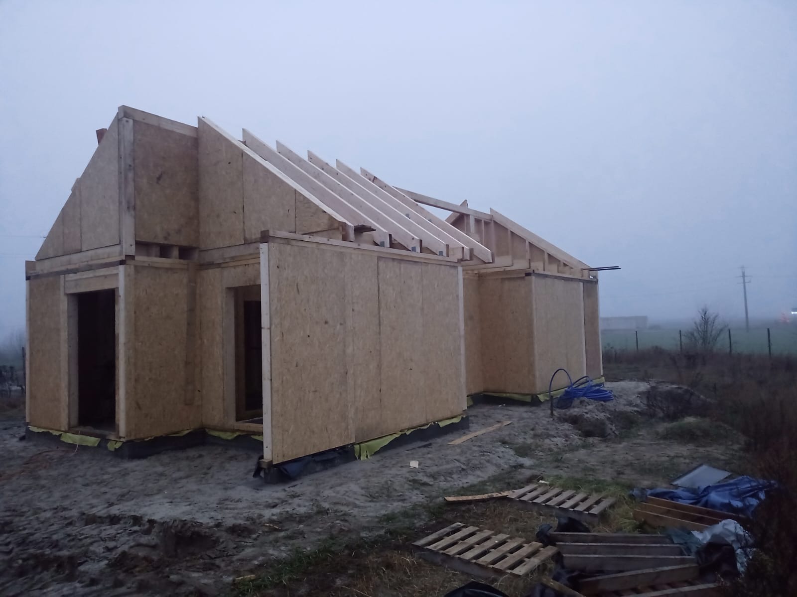 Wznoszenie domu szkieletowego: konstrukcja ścian z płyt OSB i drewnianych belek pod krokwie w pochmurny dzień. Widoczne palety i niebieski wąż na ziemi.