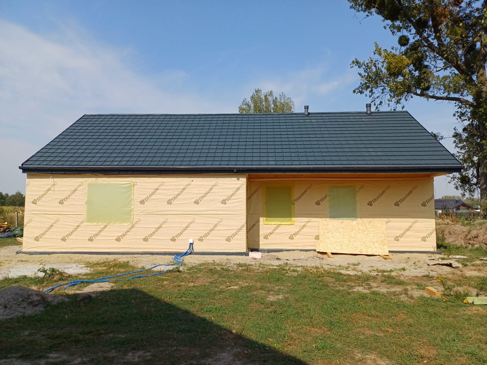 Budowa domu szkieletowego: zabezpieczona folią elewacja, grafitowa dachówka i częściowo zakryte okna, widoczne kable. Etap przed ociepleniem.