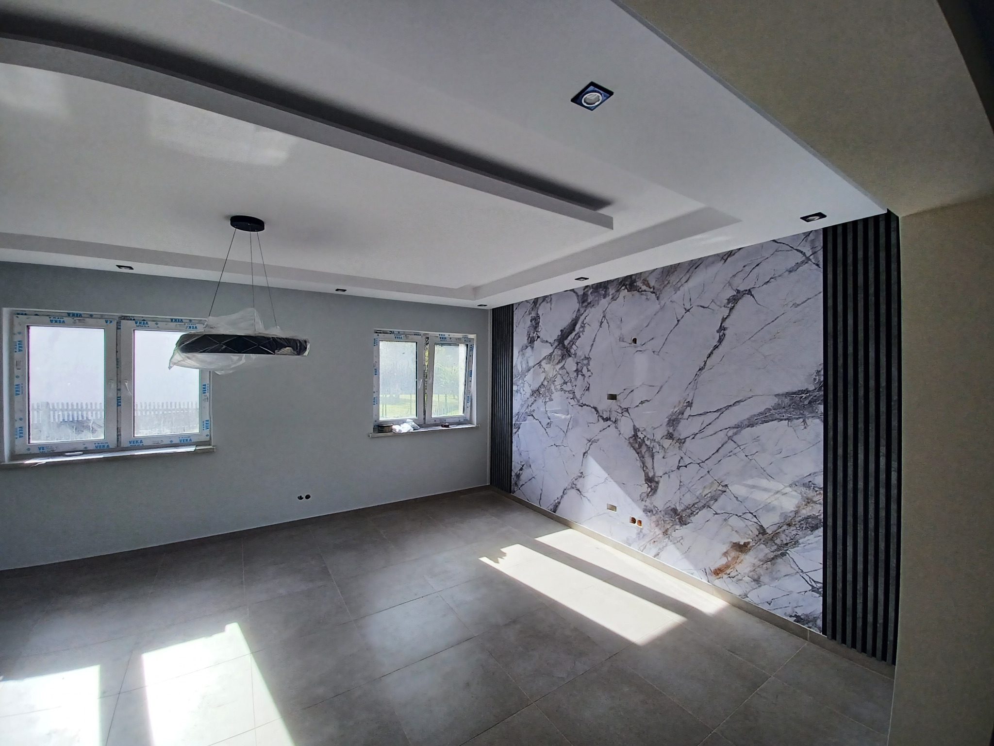Wykończone wnętrze: szare płytki podłogowe, ściana z efektem marmuru i lamele, okna PCV. Nowoczesna lampa wisząca. Całość w jasnych, chłodnych tonacjach.