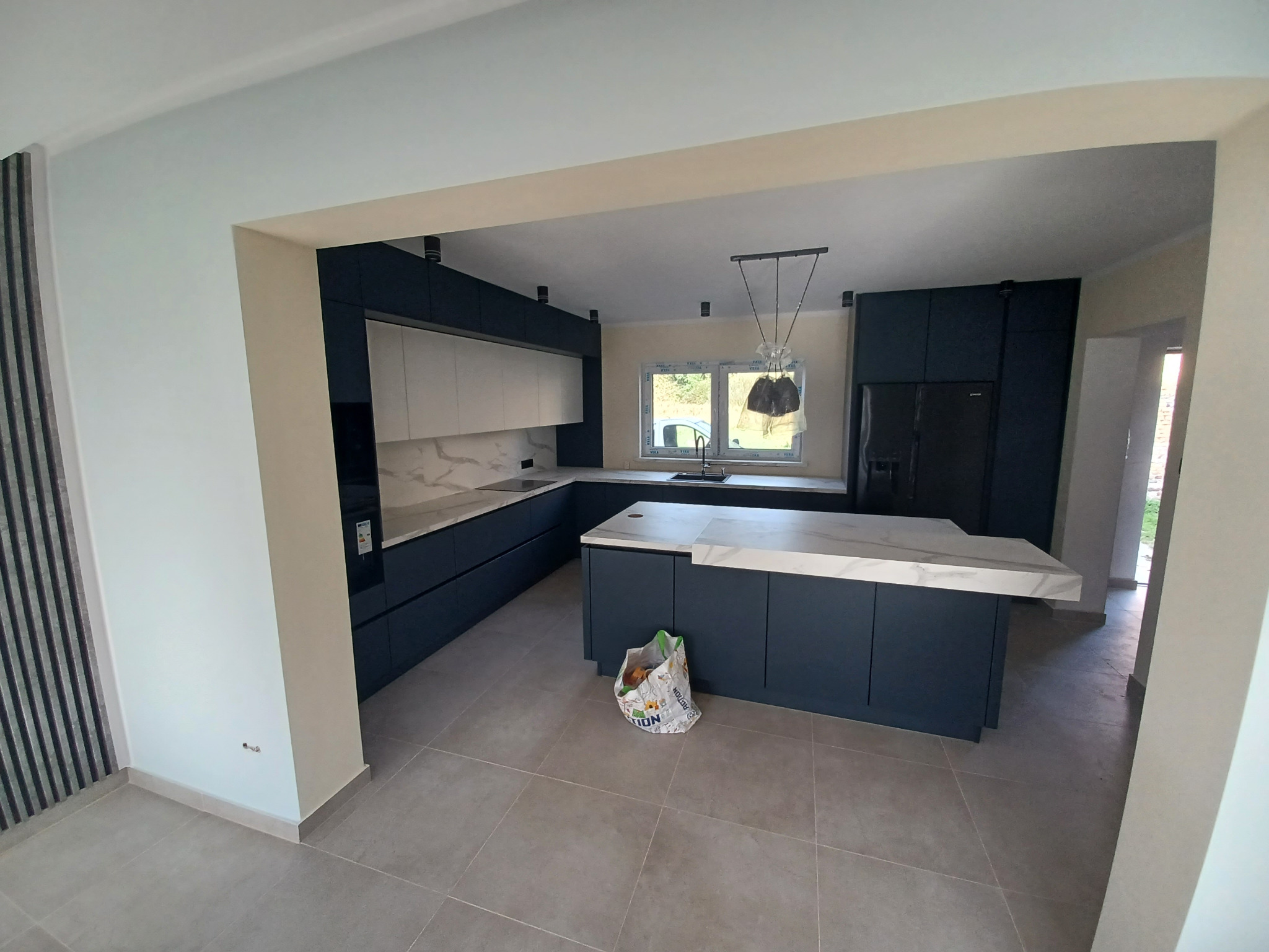 Nowoczesna kuchnia z wyspą i granatowymi szafkami, wykończona marmurowym blatem i płytkami podłogowymi. Widoczne detale oświetlenia i dekoracyjne panele ścienne.