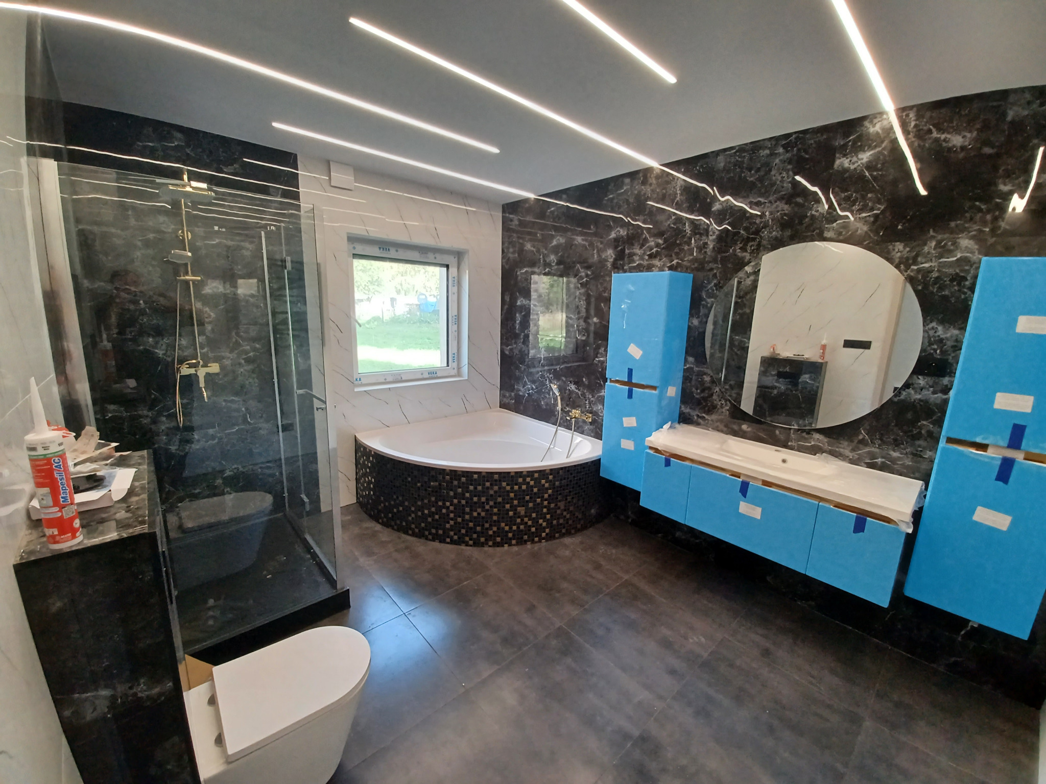 Nowoczesna łazienka z czarnym marmurem i podświetlanym sufitem. Kabina prysznicowa ze złotą armaturą, wanna narożna z mozaiką i niebieskie szafki pod umywalkę.