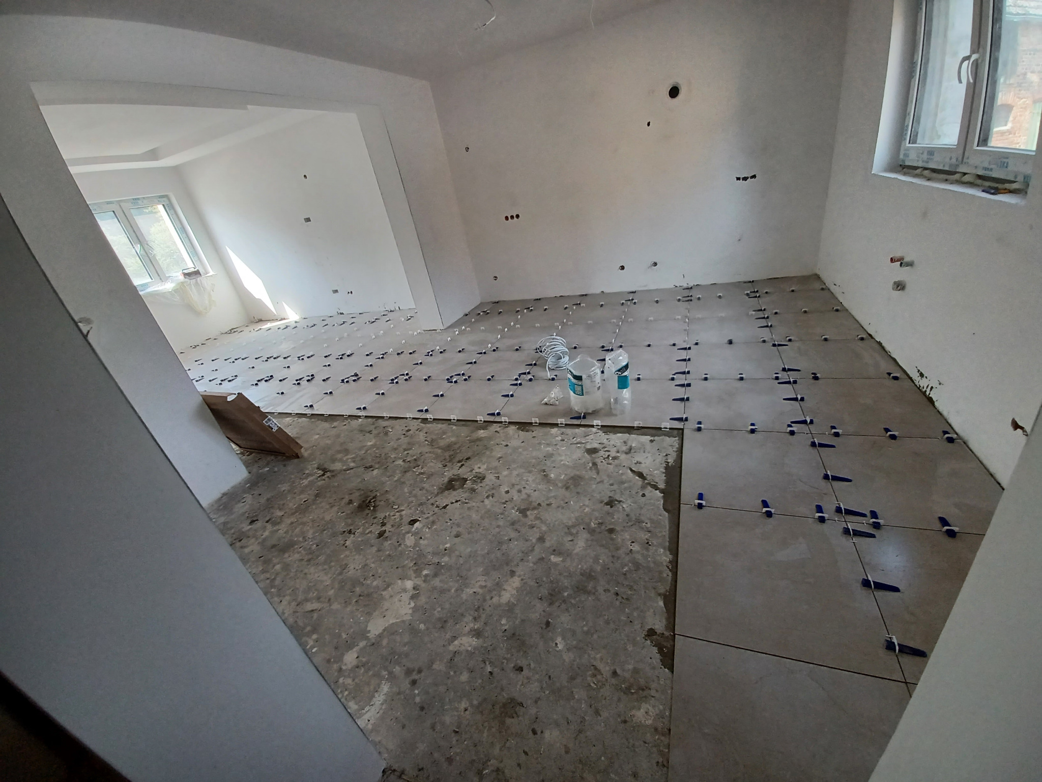 Układanie płytek podłogowych w jasnym pomieszczeniu. Widoczne krzyżyki dystansowe i częściowo ułożona podłoga. Fragment surowej posadzki w lewym dolnym rogu.