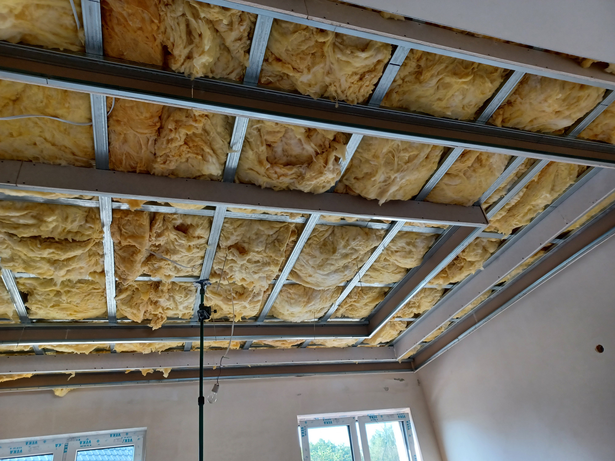 Sufit podwieszany z izolacją z wełny mineralnej, widok z dołu. Metalowa konstrukcja kratowa. W tle okno i ściana w surowym tynku.