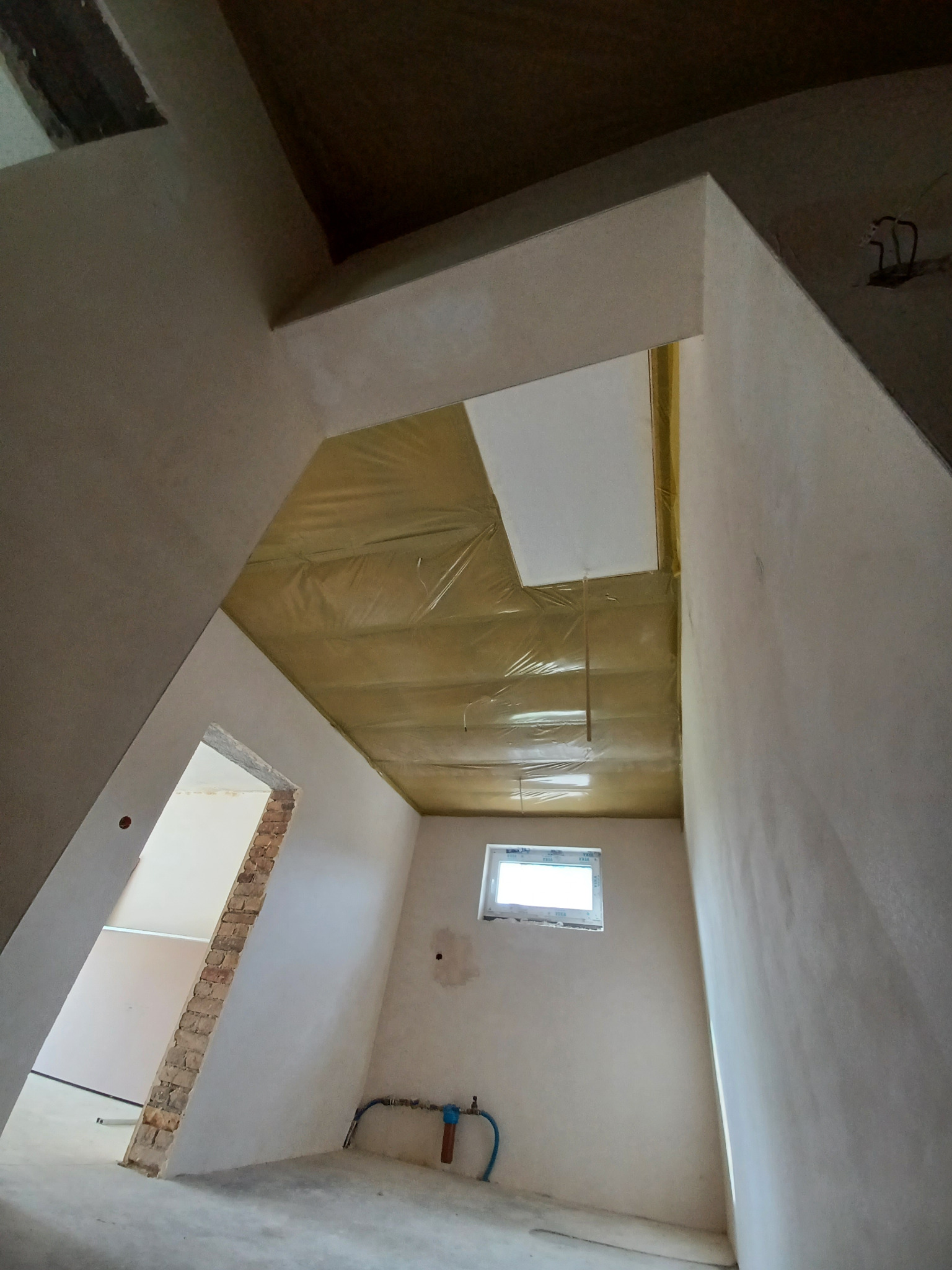 Surowe wnętrze poddasza z oknem, izolacją stropu i ścianami po gładziach, przygotowane do dalszych prac wykończeniowych. Widok z dołu.