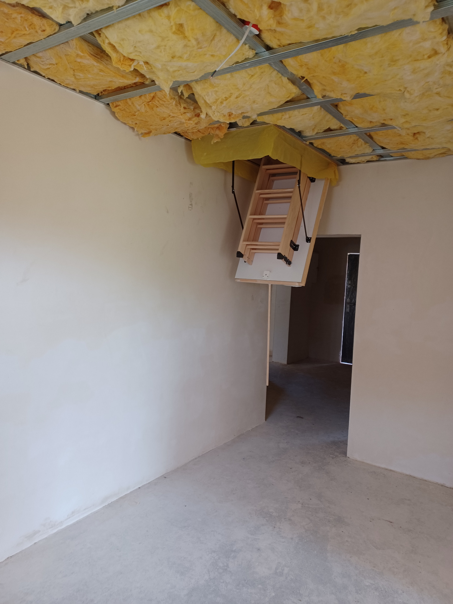 Otwierane drewniane schody strychowe z żółtą izolacją termiczną na suficie podwieszanym w surowym, jasnym pomieszczeniu w trakcie wykańczania.