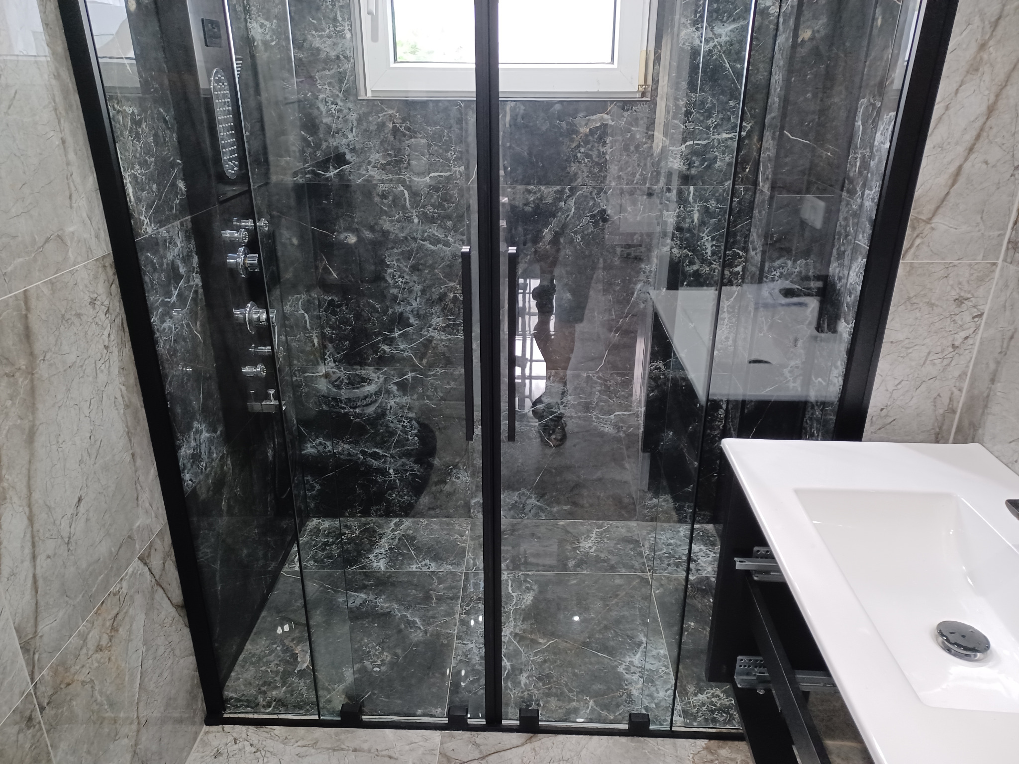 Nowoczesna kabina prysznicowa z czarnymi profilami i marmurowymi płytkami. Widoczna biała umywalka. Całość utrzymana w eleganckiej, minimalistycznej stylistyce.