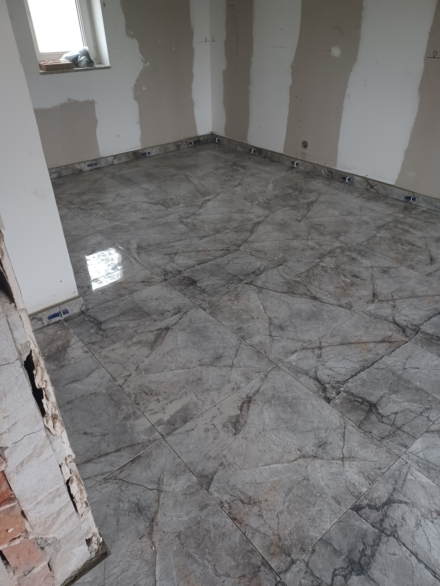 Wykończona podłoga z szarych płytek imitujących marmur, z niebieskimi krzyżykami dystansowymi przy ścianach, w trakcie prac wykończeniowych pomieszczenia.