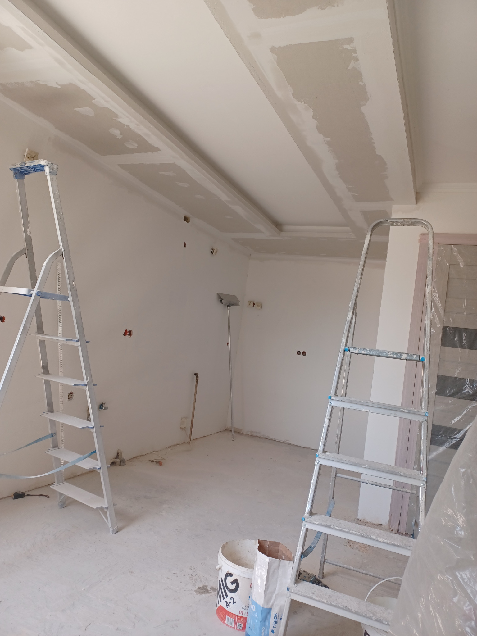 Wnętrze w trakcie prac wykończeniowych: widoczne dwie drabiny, wiadro z gładzią i częściowo wykończony sufit z płyt kartonowo-gipsowych.