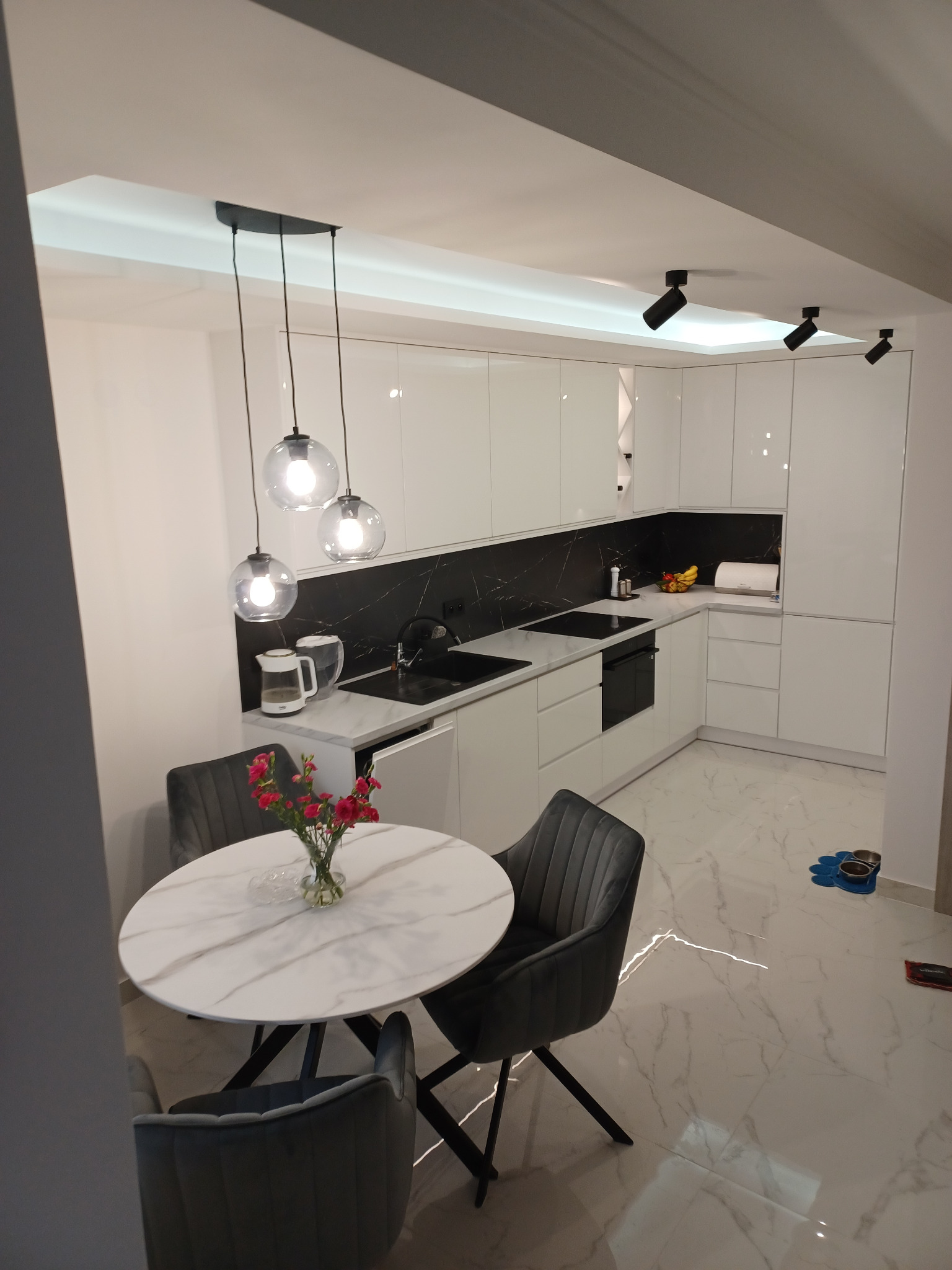 Nowoczesna, biała kuchnia z czarnym blatem i marmurową podłogą. Okrągły stół z szarymi krzesłami i kwiatami. Wiszące lampy kuliste i punktowe oświetlenie sufitowe.