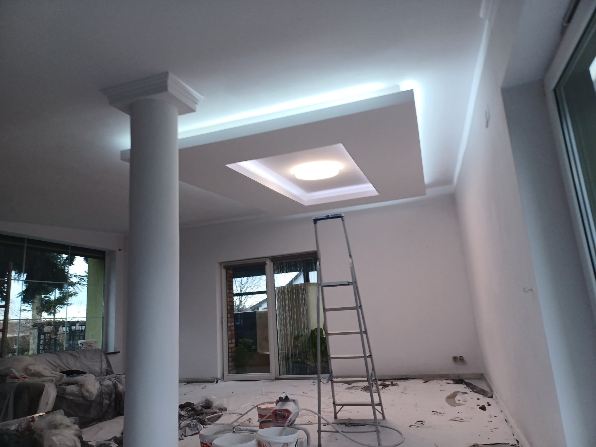 Wnętrze w trakcie prac wykończeniowych: kolumna, sufit z oświetleniem LED, drabina malarska, okna. Częściowo zabezpieczone meble. Widoczne ślady farby.