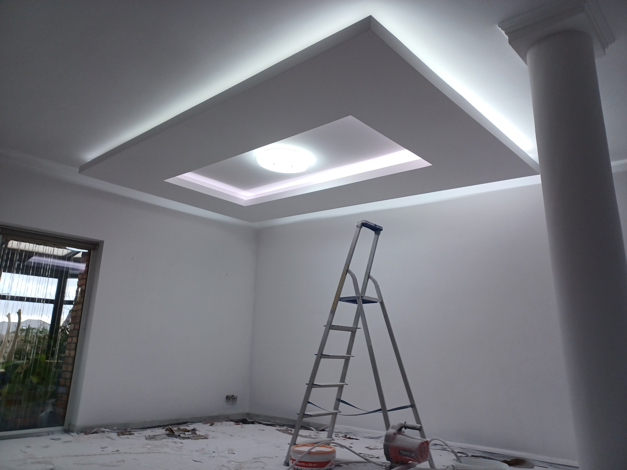 Oświetlenie sufitowe z płyt karton-gips w kształcie kwadratu z kolumną. Widoczna drabina malarska i resztki po remoncie na podłodze.