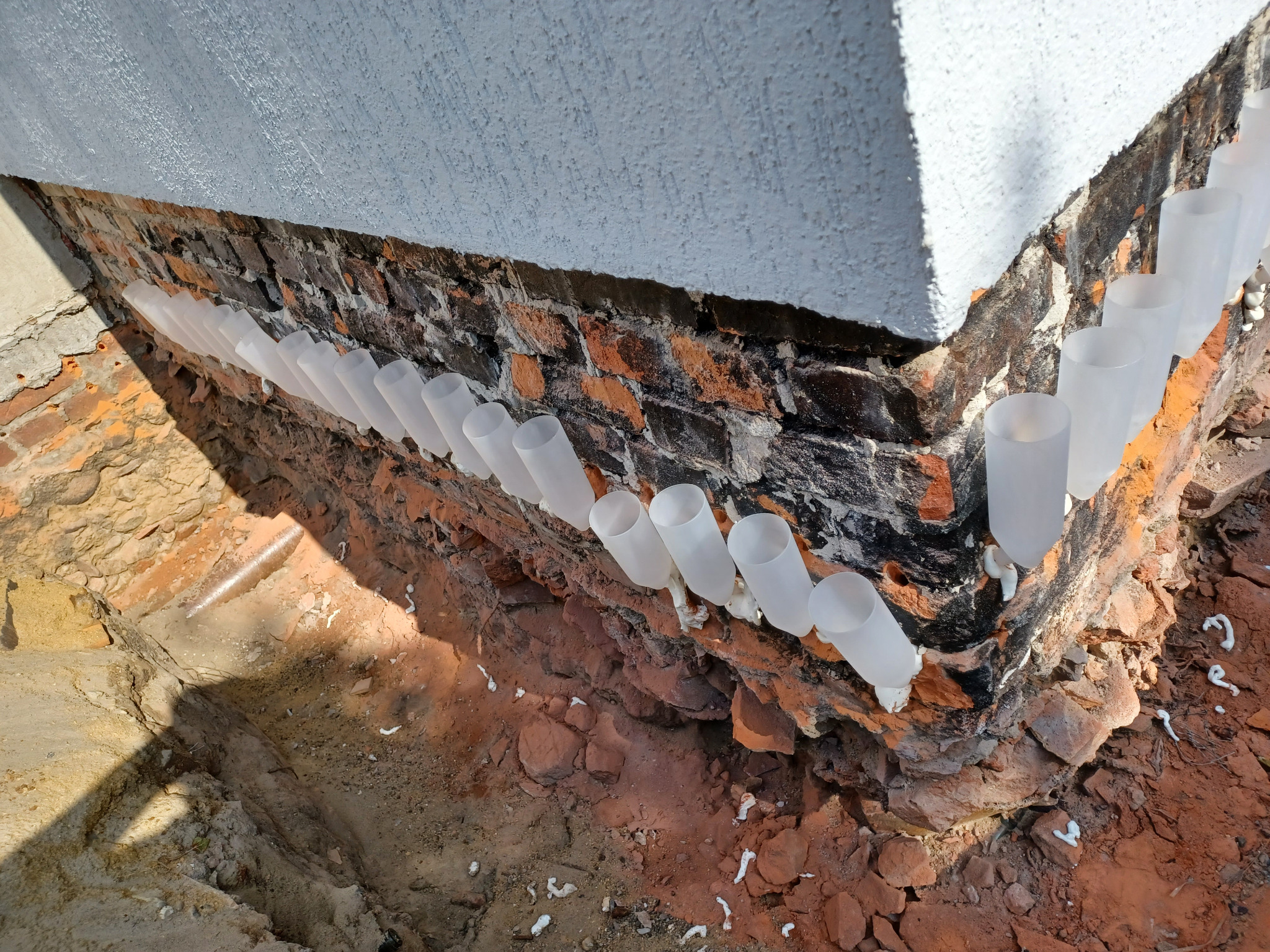 Odsłonięty fundament budynku z cegły, zabezpieczony przezroczystymi rurkami z pianką montażową, w wykopie ziemnym. Izolacja przeciwwilgociowa.