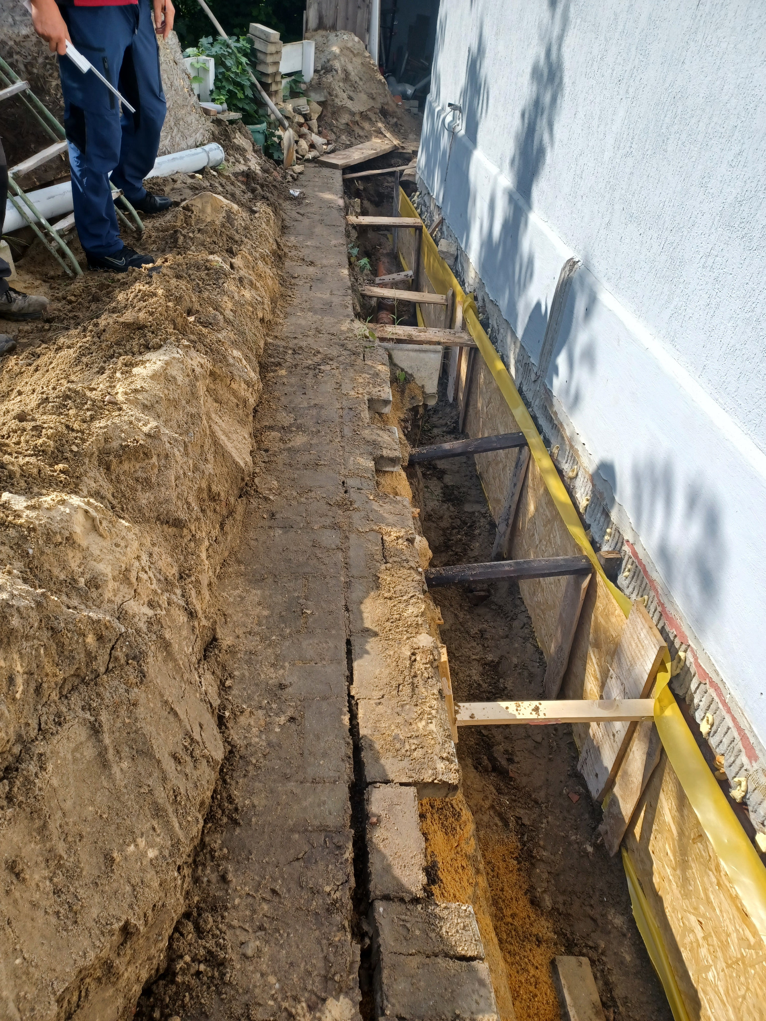 Wykop fundamentów z drewnianą szalunką przy ścianie budynku, przygotowany do wylania betonu. Widoczny fragment ubrudzonych ziemią spodni i drabina.