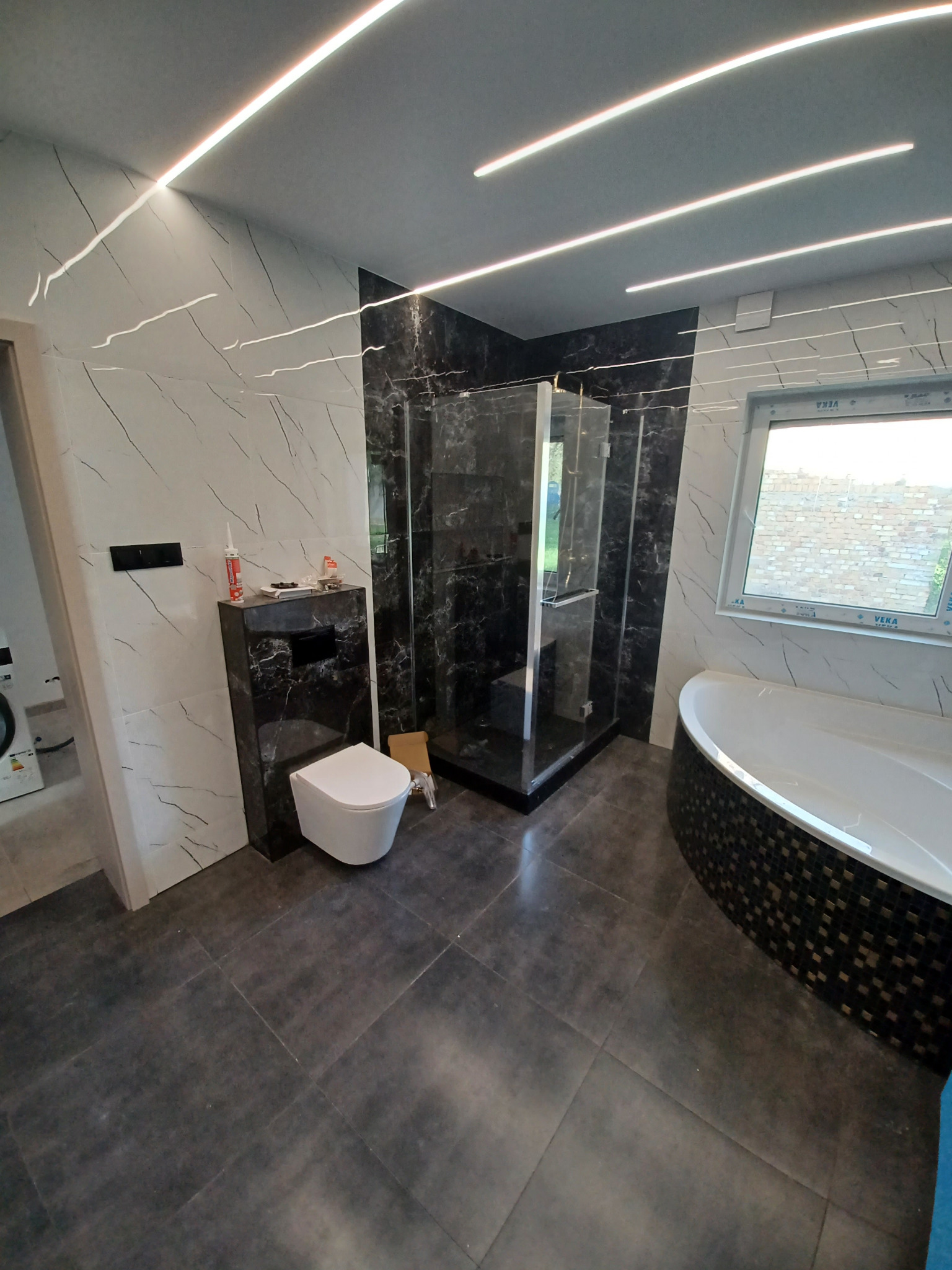 Nowoczesna łazienka z kabiną prysznicową typu walk-in, podświetleniem LED i marmurowymi płytkami. Uwagę zwraca nietypowa wanna z mozaiką.