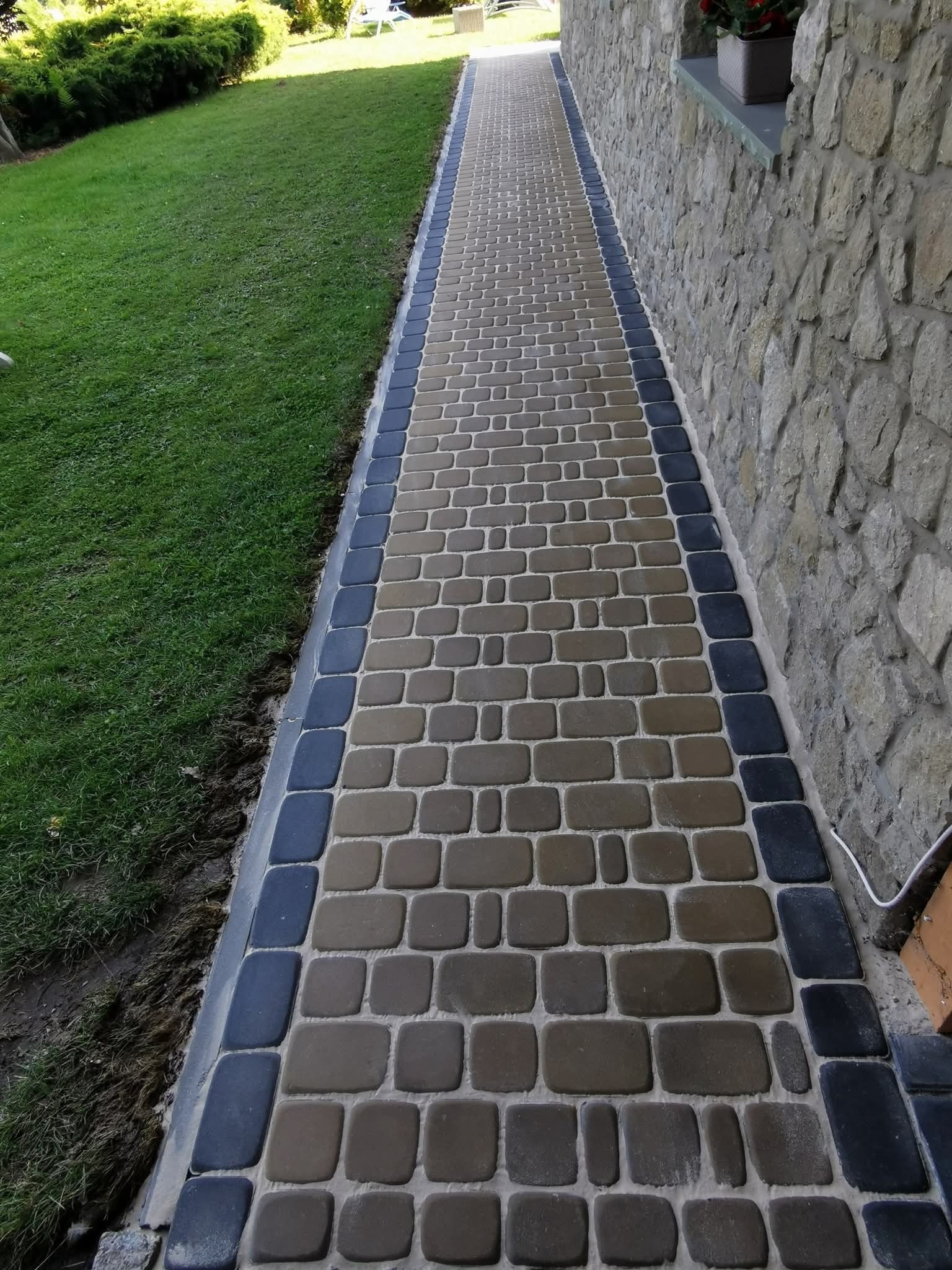 Długa ścieżka z kostki brukowej w odcieniach brązu i grafitu, biegnąca wzdłuż kamiennej ściany i trawnika.  Widok z perspektywy.