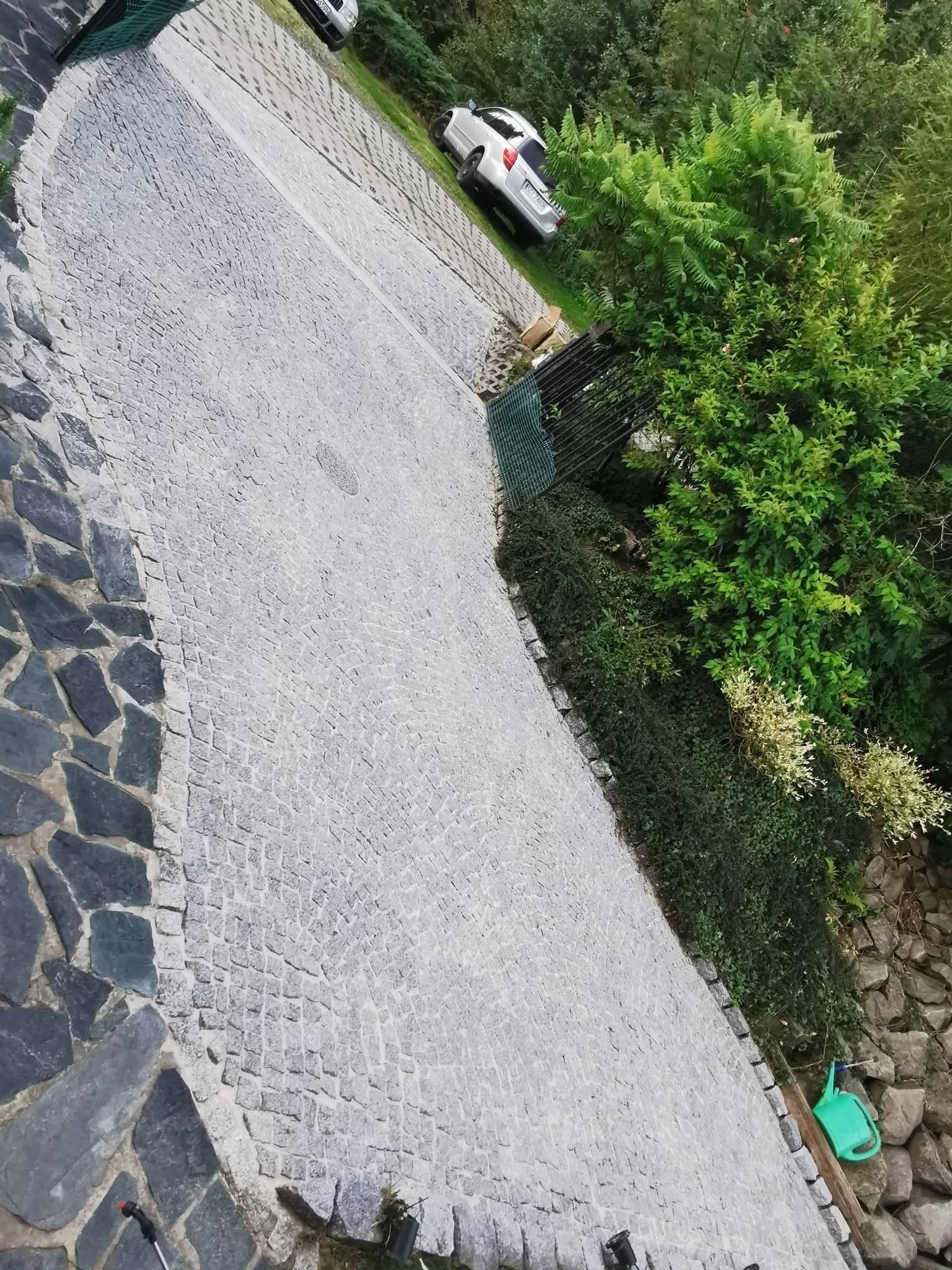 Szara kostka brukowa na podjeździe, widok z góry, obok zieleń i zaparkowany samochód. Widoczne narzędzia ogrodowe. Ujęcie z bliska, podkreślające fakturę kostki.