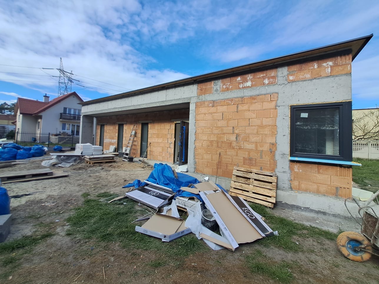 Wznoszenie parterowego domu z cegły ceramicznej, z oknem w czarnej ramie. Widoczne materiały budowlane, drabina i częściowo wykończony dach.