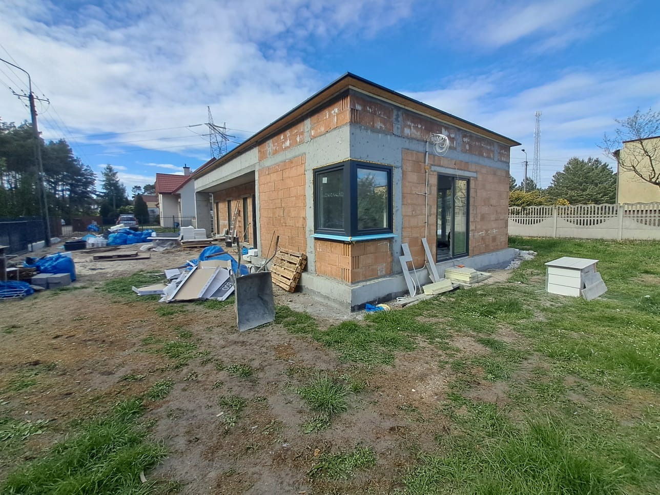 Budowa domu z cegły ceramicznej: ściany w trakcie wznoszenia, okna wstawione, widoczne materiały budowlane na zewnątrz. Stan surowy otwarty, widok na elewację boczną.