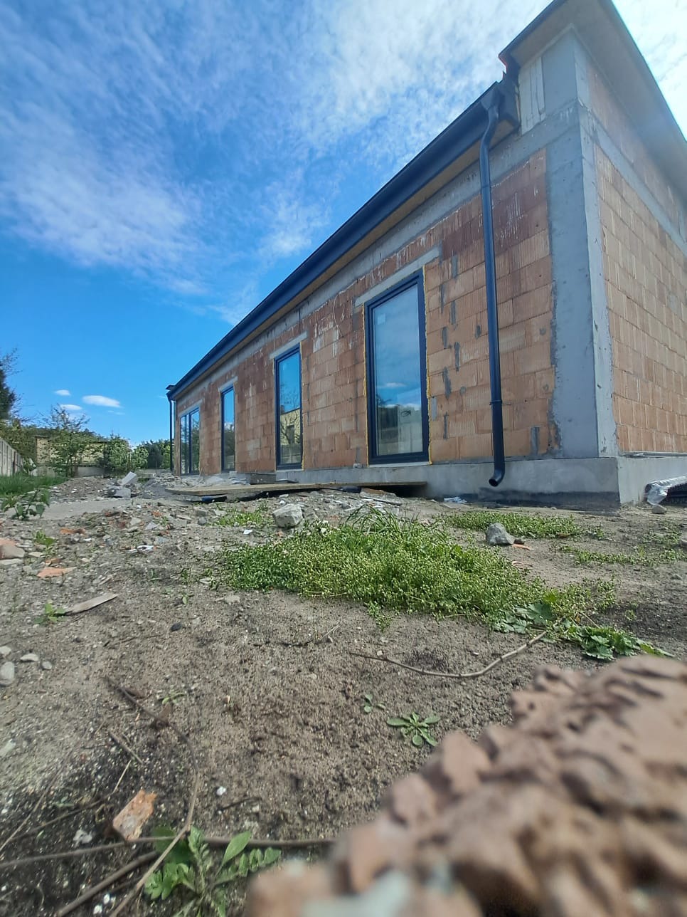 Niedokończony murowany dom z czarnymi oknami i rynnami, widoczny z niskiej perspektywy na tle błękitnego nieba. Surowy stan budynku, teren wokół w trakcie przygotowania.