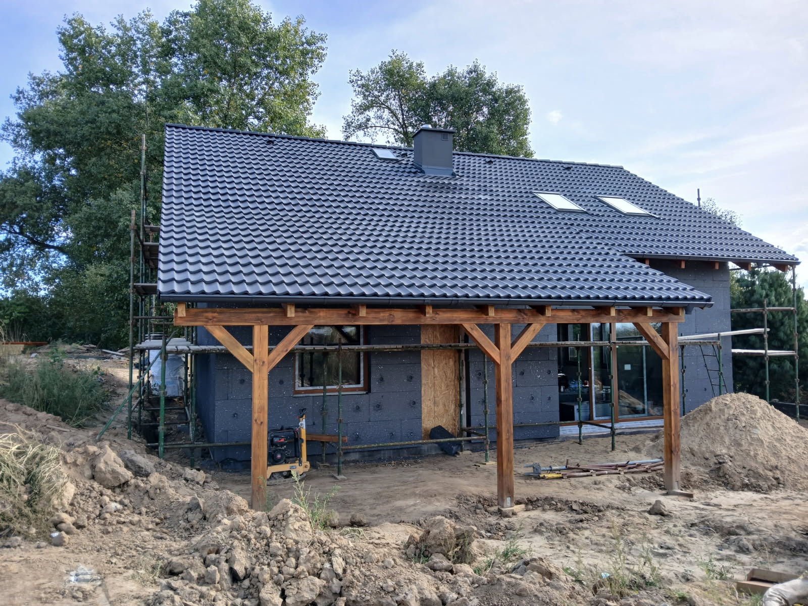 Dom w trakcie budowy: szara elewacja ocieplona styropianem, grafitowy dach z dachówką, drewniany podcień, rusztowania, ziemia. Widok z zewnątrz, etap prac.