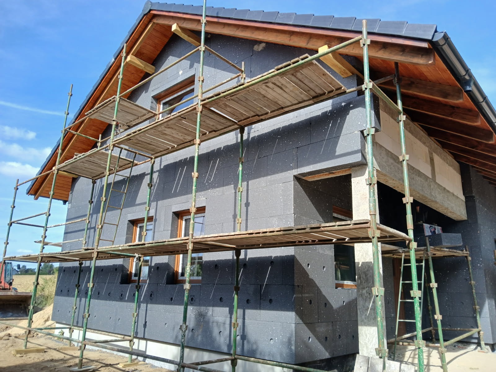 Budowa domu: ściany z izolacją termiczną, rusztowania. Widoczne okna i fragment dachu. Na drugim planie koparka i niebo.