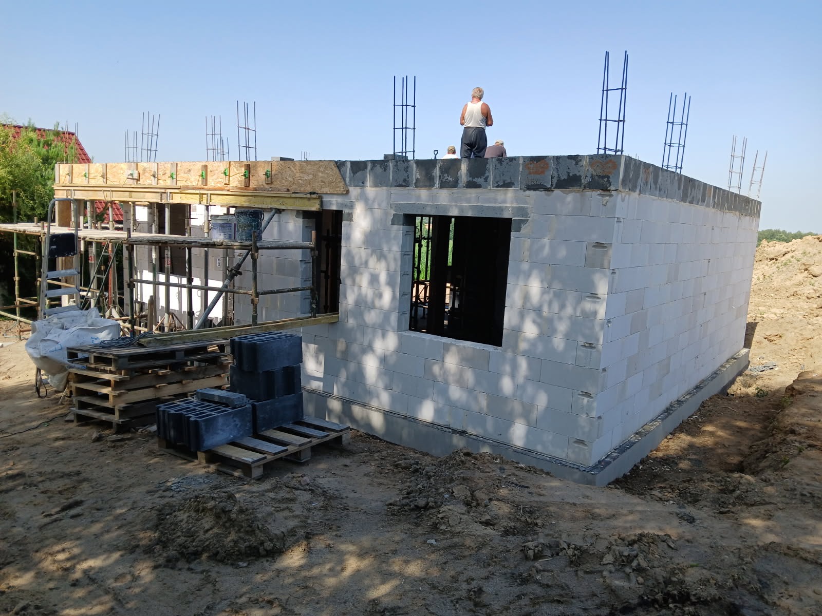 Budowa domu z białych bloczków, widoczne zbrojenia, szalunki okienne i rusztowania. Na dachu pracownicy budowlani. Wokół plac budowy i palety z bloczkami.