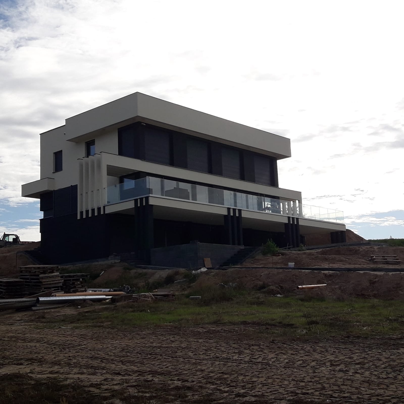Nowoczesny, dwukondygnacyjny dom z płaskim dachem i szklaną balustradą na balkonie. Widoczne elementy konstrukcyjne i teren wokół w trakcie prac wykończeniowych.