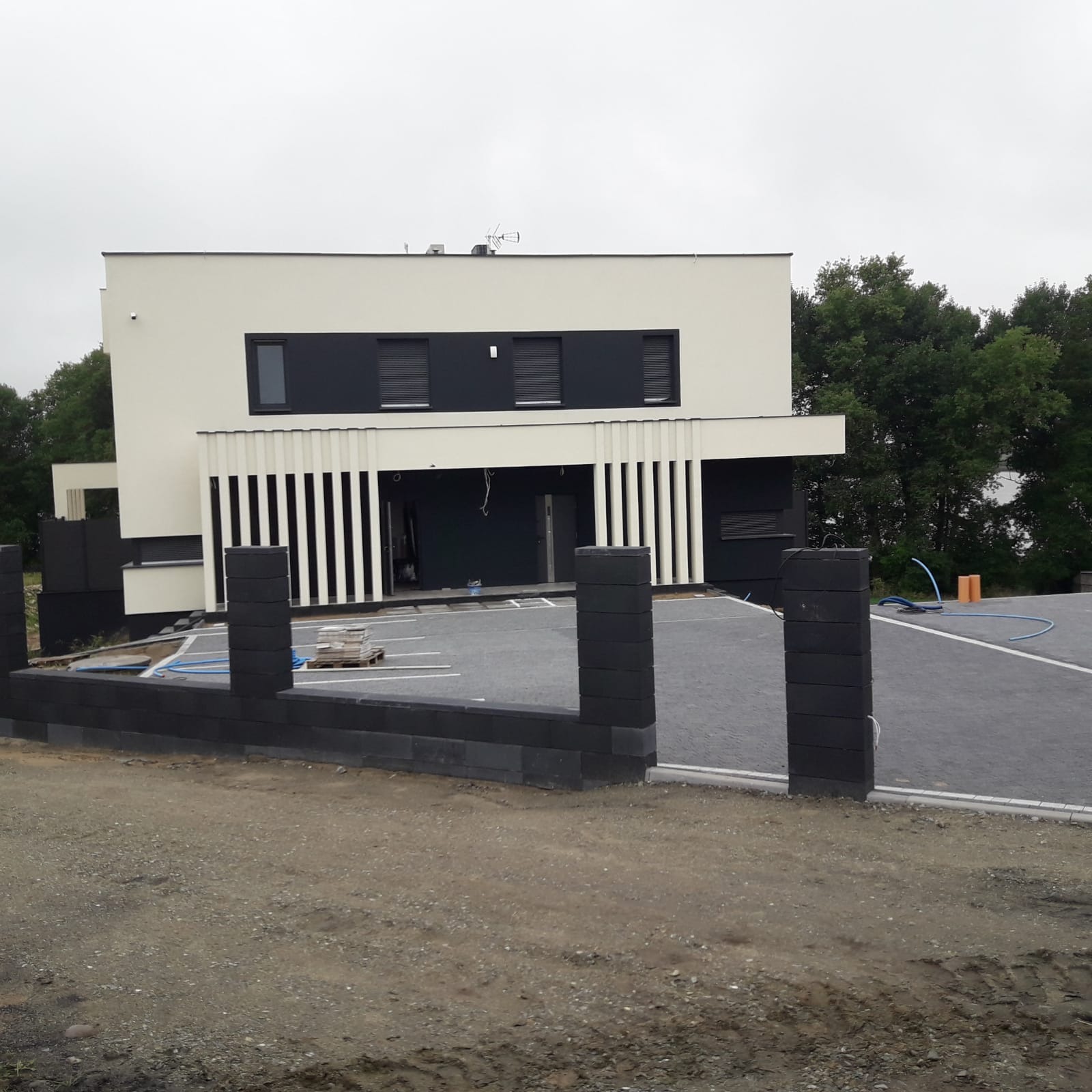 Nowoczesny dom z płaskim dachem i elementami dekoracyjnymi. Ułożona kostka brukowa na podjeździe, w tle drzewa. Częściowo ukończone ogrodzenie z szarych bloczków.