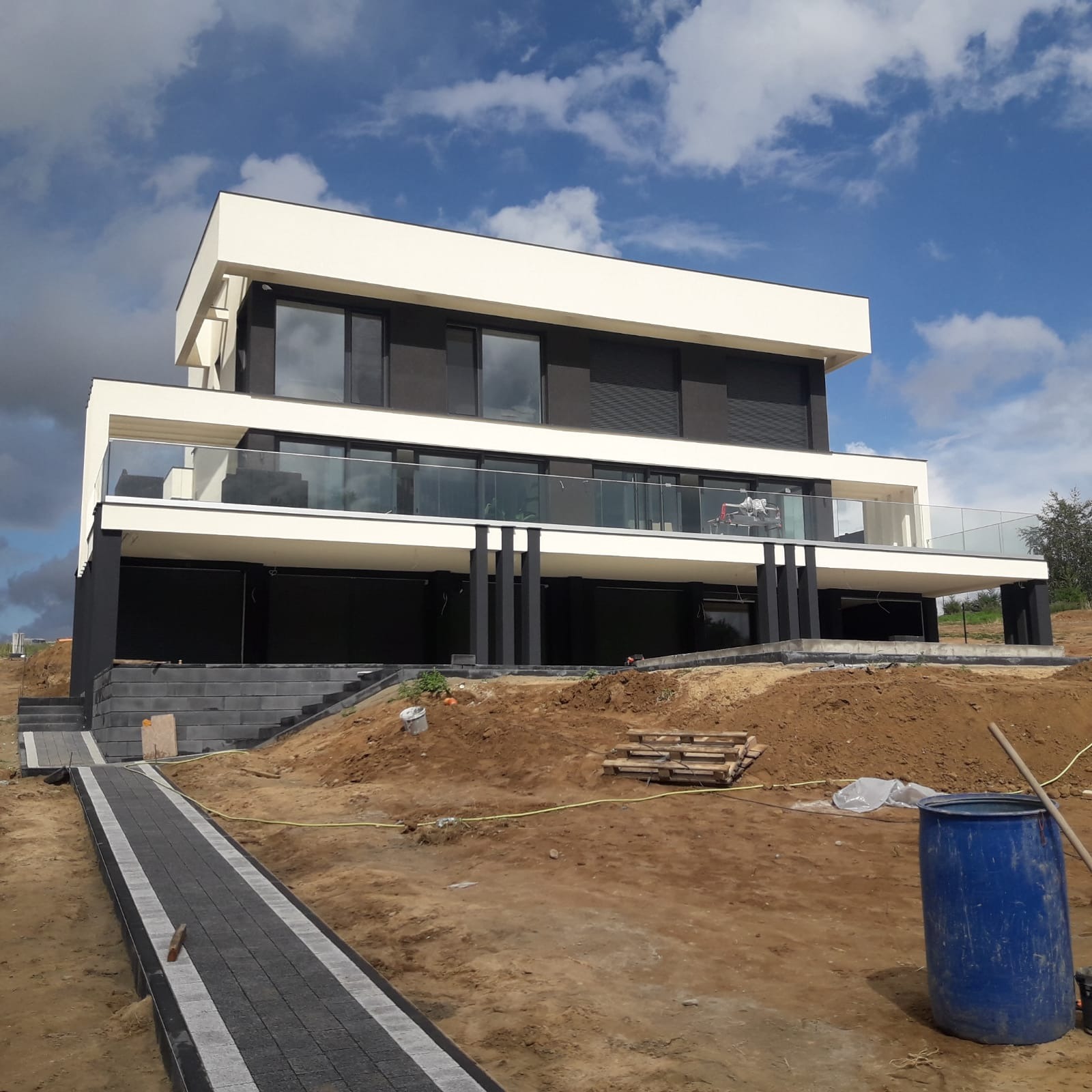 Nowoczesny, dwupiętrowy dom z płaskim dachem w trakcie budowy. Widoczne szare ścieżki prowadzące do budynku, niebieska beczka i palety na ziemi.