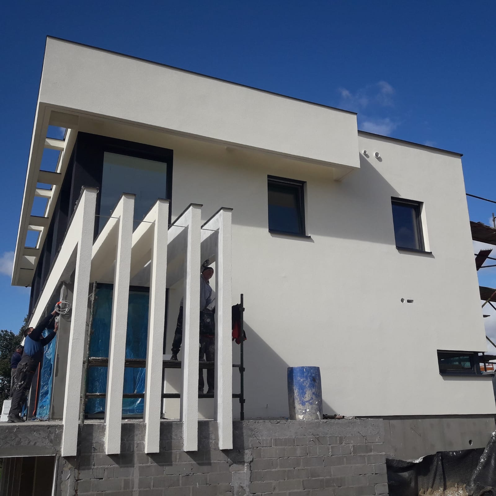 Nowoczesny dom z białym wykończeniem elewacji w trakcie prac malarskich. Widoczni pracownicy na rusztowaniu i drabina. Błękitne niebo w tle, częściowo zakryte przez budynek.