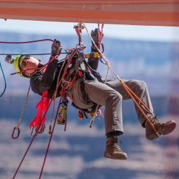 ProALPINISTA Kyrylo Rozhentsov - Osoba w uprzęży, kasku i okularach, w trakcie pracy na wysokości, zawieszona na linach z widokiem na rozmyte tło skalne i błękitne niebo.