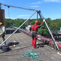 ProALPINISTA Kyrylo Rozhentsov - Prace wysokościowe na dachu budynku z użyciem specjalistycznego sprzętu asekuracyjnego. Dwóch pracowników w kaskach i kombinezonach zabezpieczających.