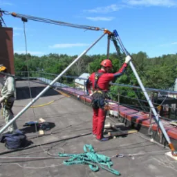 Prace wysokościowe na dachu budynku z użyciem specjalistycznego sprzętu asekuracyjnego. Dwóch pracowników w kaskach i kombinezonach zabezpieczających.