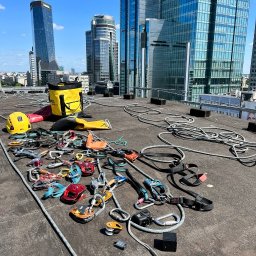 ProALPINISTA Kyrylo Rozhentsov - Sprzęt do prac wysokościowych rozłożony na dachu z widokiem na panoramę miasta. Żółty worek, kaski, liny i karabinki tworzą przygotowanie do zadania.