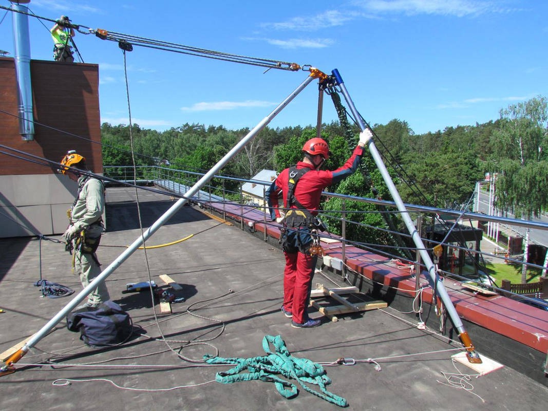 Prace wysokościowe na dachu budynku z użyciem specjalistycznego sprzętu asekuracyjnego. Dwóch pracowników w kaskach i kombinezonach zabezpieczających.