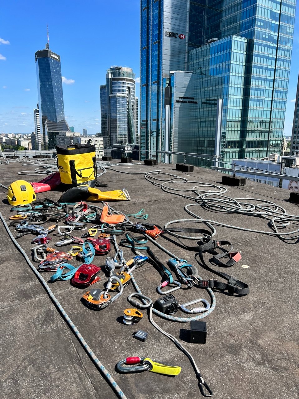 Sprzęt do prac wysokościowych rozłożony na dachu z widokiem na panoramę miasta. Żółty worek, kaski, liny i karabinki tworzą przygotowanie do zadania.