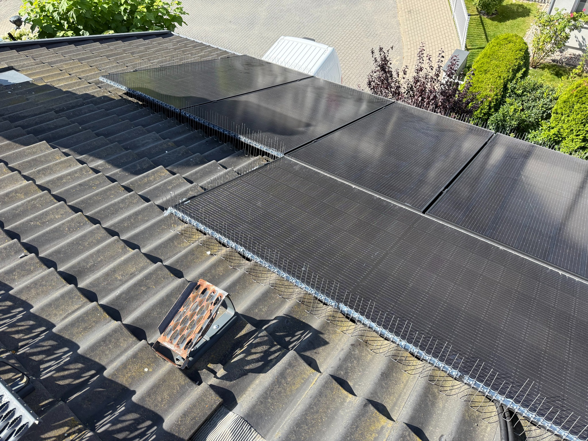 Ukończony montaż paneli fotowoltaicznych z zabezpieczeniem przed ptakami na dachu pokrytym dachówką. Widoczny fragment ogrodu i chodnika.