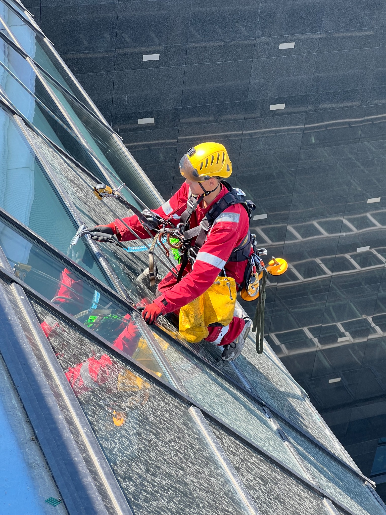 Pracownik w czerwonym kombinezonie, z żółtym kaskiem, myje okna na wysokościowcu, używając ściągaczki. Widoczne liny, sprzęt asekuracyjny i odblask w szybach.