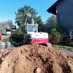 PNEUMATIC HYDRO - Mini koparka TB290 na tle domu z ociepleniem, w trakcie prac ziemnych. Widoczny wykop, ziemia i osoba w czerwonej koszulce. Słoneczny dzień.