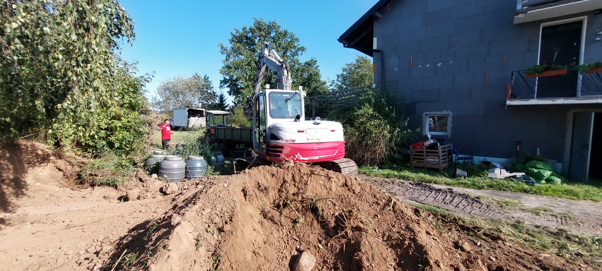 Mini koparka TB290 na tle domu z ociepleniem, w trakcie prac ziemnych. Widoczny wykop, ziemia i osoba w czerwonej koszulce. Słoneczny dzień.