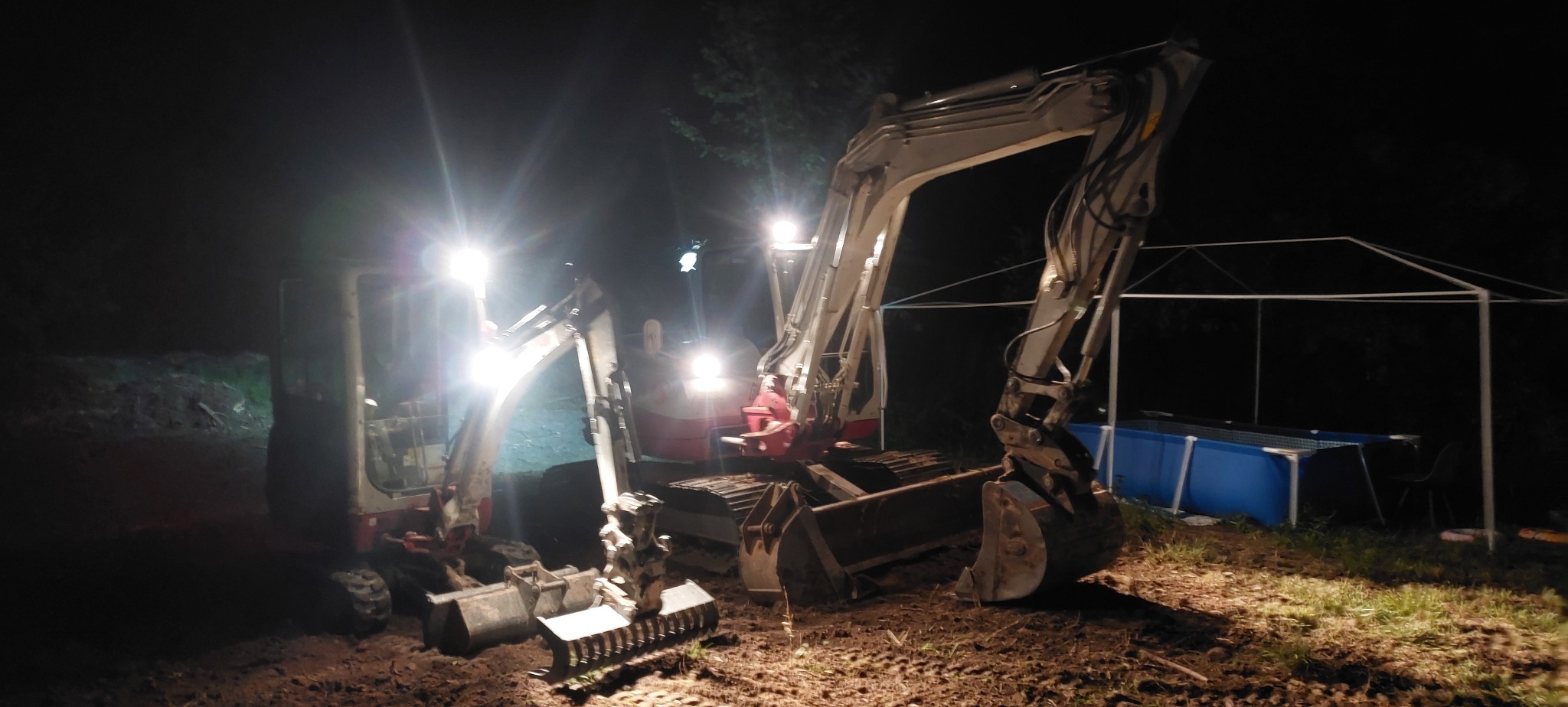 Dwa oświetlone minikoparki na placu budowy nocą, z widocznym basenem stelażowym w tle. Jeden z osprzętów leży na ziemi. Scena robót ziemnych po zmroku.