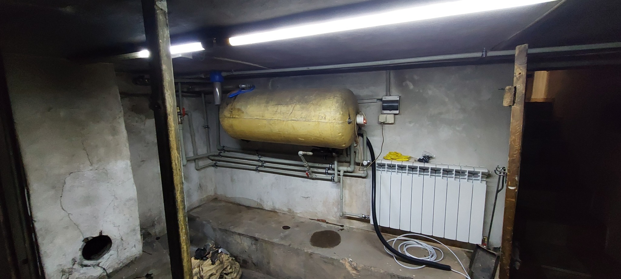 Żółty zbiornik hydroforowy w piwnicy, widoczne rury, grzejnik, przewody i narzędzia. Ściany surowe, oświetlenie jarzeniowe.