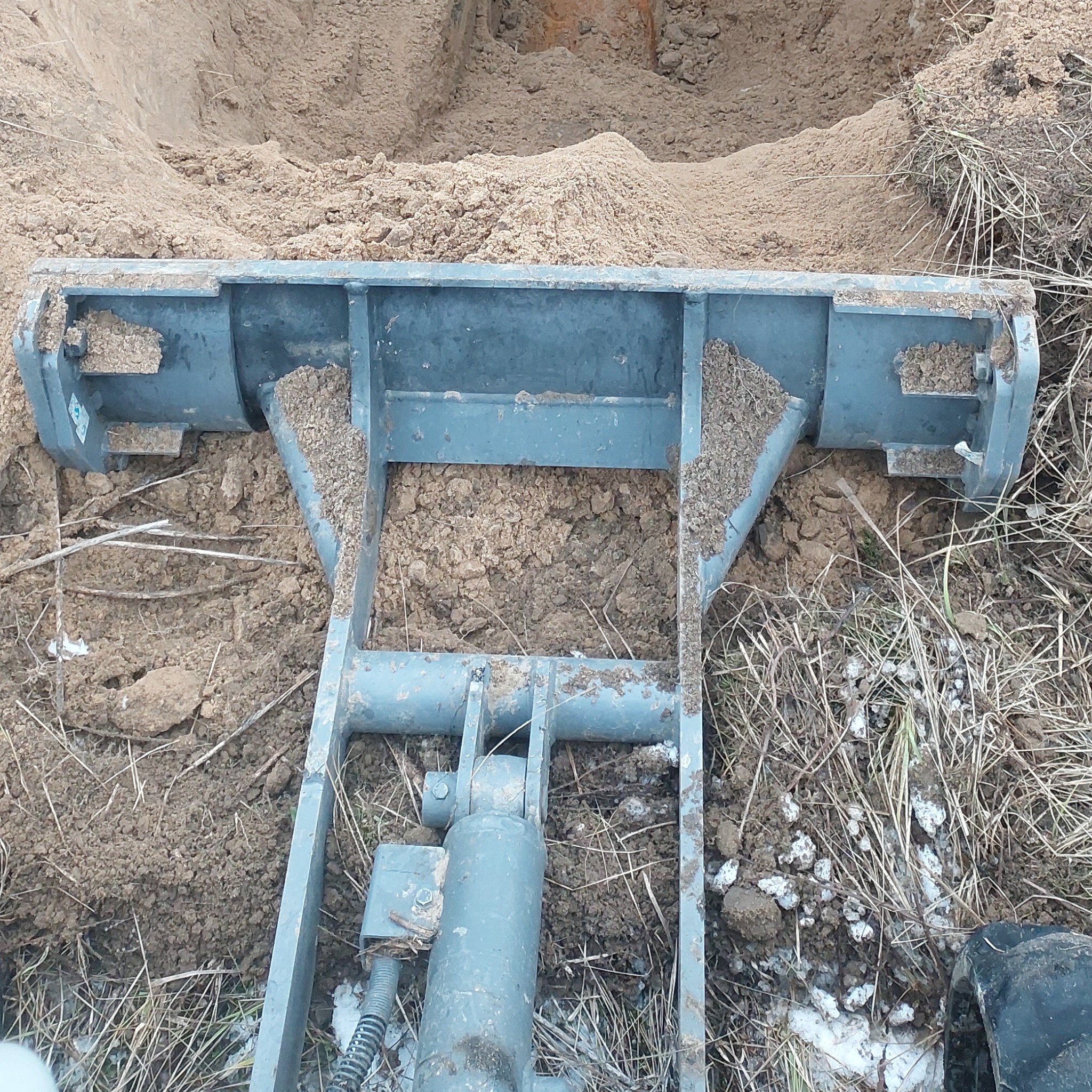 Szczegółowe ujęcie łyżki koparki zagłębiającej się w ziemię, widoczne ślady ziemi na metalowej konstrukcji, częściowo ośnieżony teren.