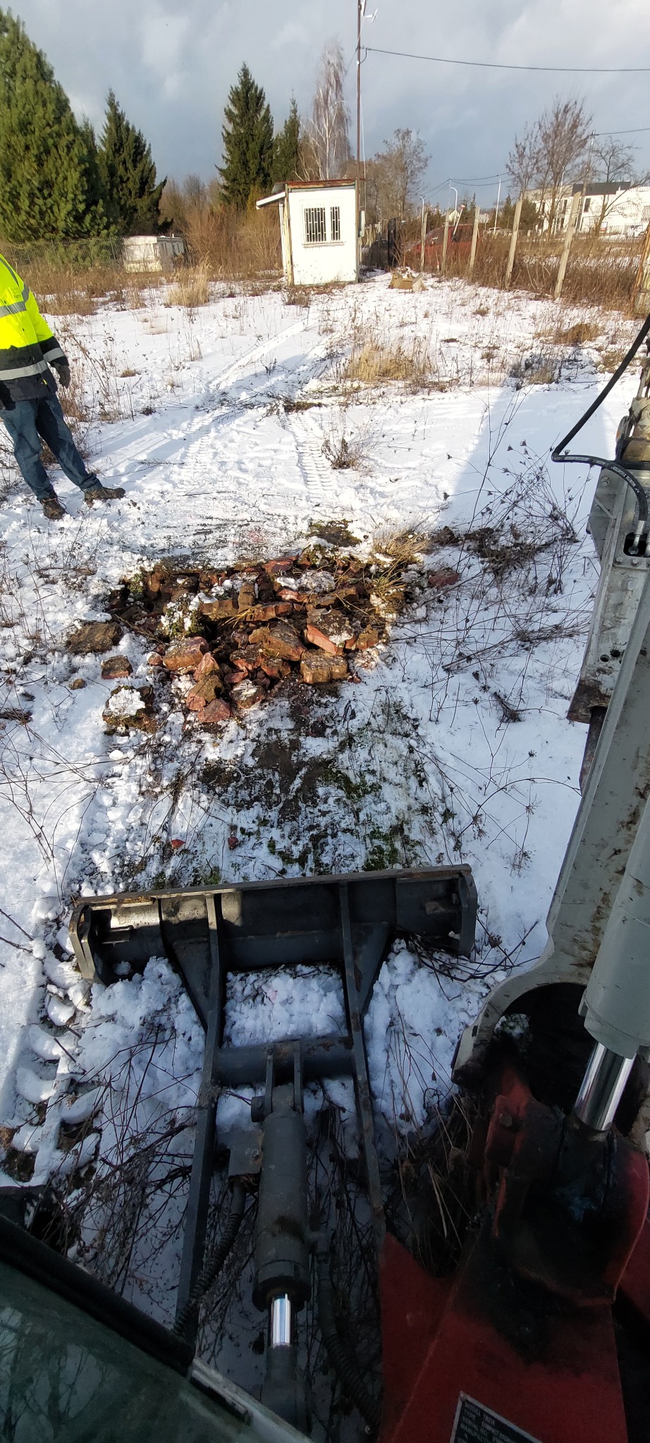 Zimowy krajobraz z wykopem gruzu i fragmentem maszyny budowlanej. W tle widoczny pracownik w odblaskowej kamizelce i mały budynek.