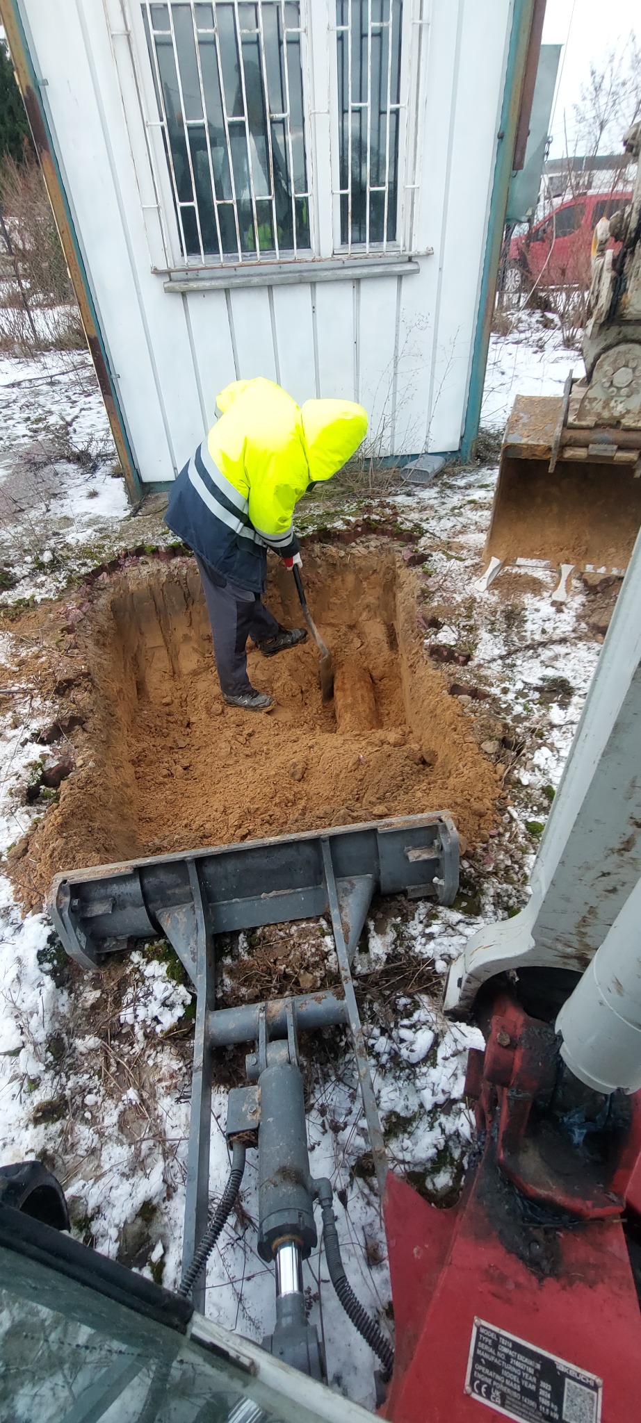 Pracownik w odblaskowej kurtce kopie dół łopatą obok białego baraku. Widoczny fragment koparki i resztki śniegu na ziemi. Ujęcie z perspektywy operatora maszyny.