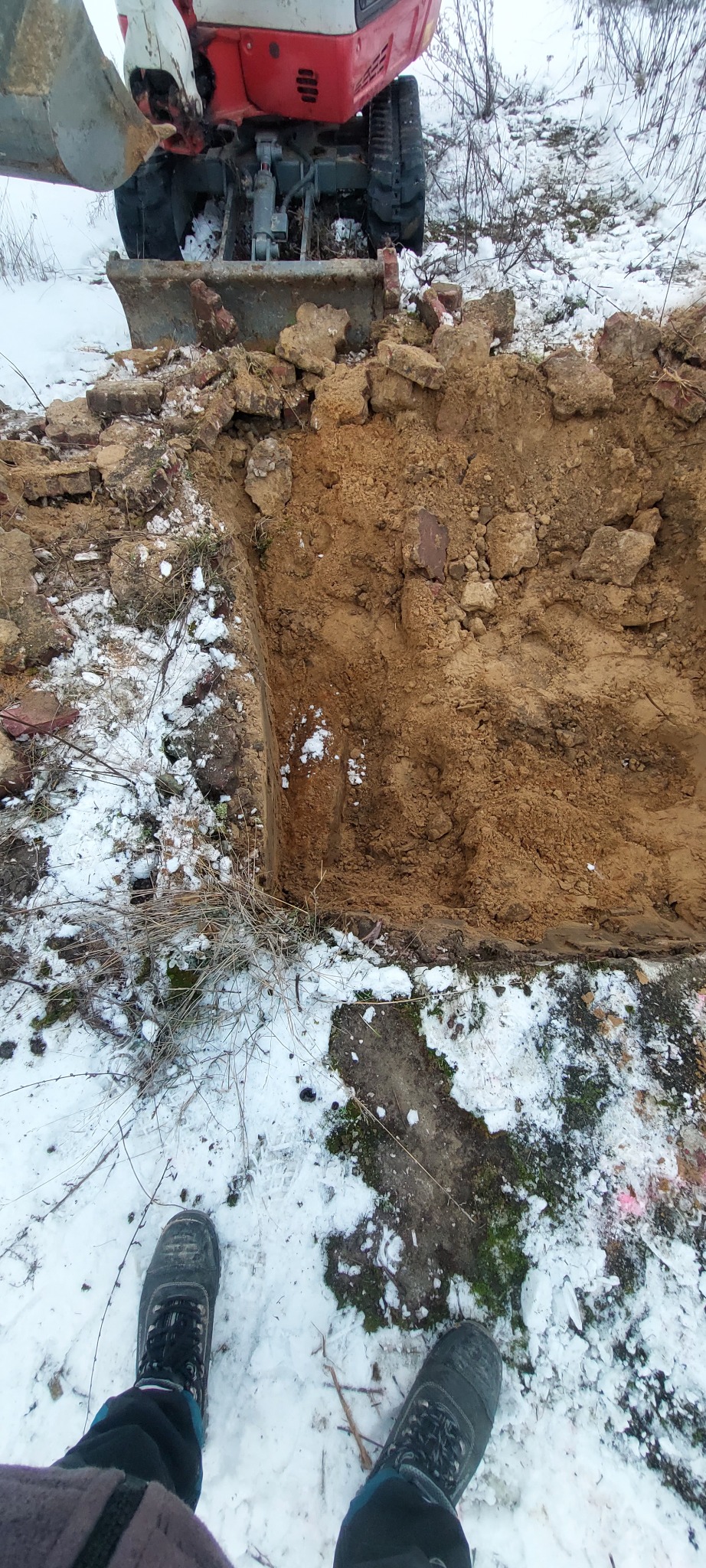 Mini koparka wykonuje wykop w ziemi, widoczne ślady śniegu. Ujęcie z perspektywy osoby stojącej obok wykopu, w tle fragment maszyny.