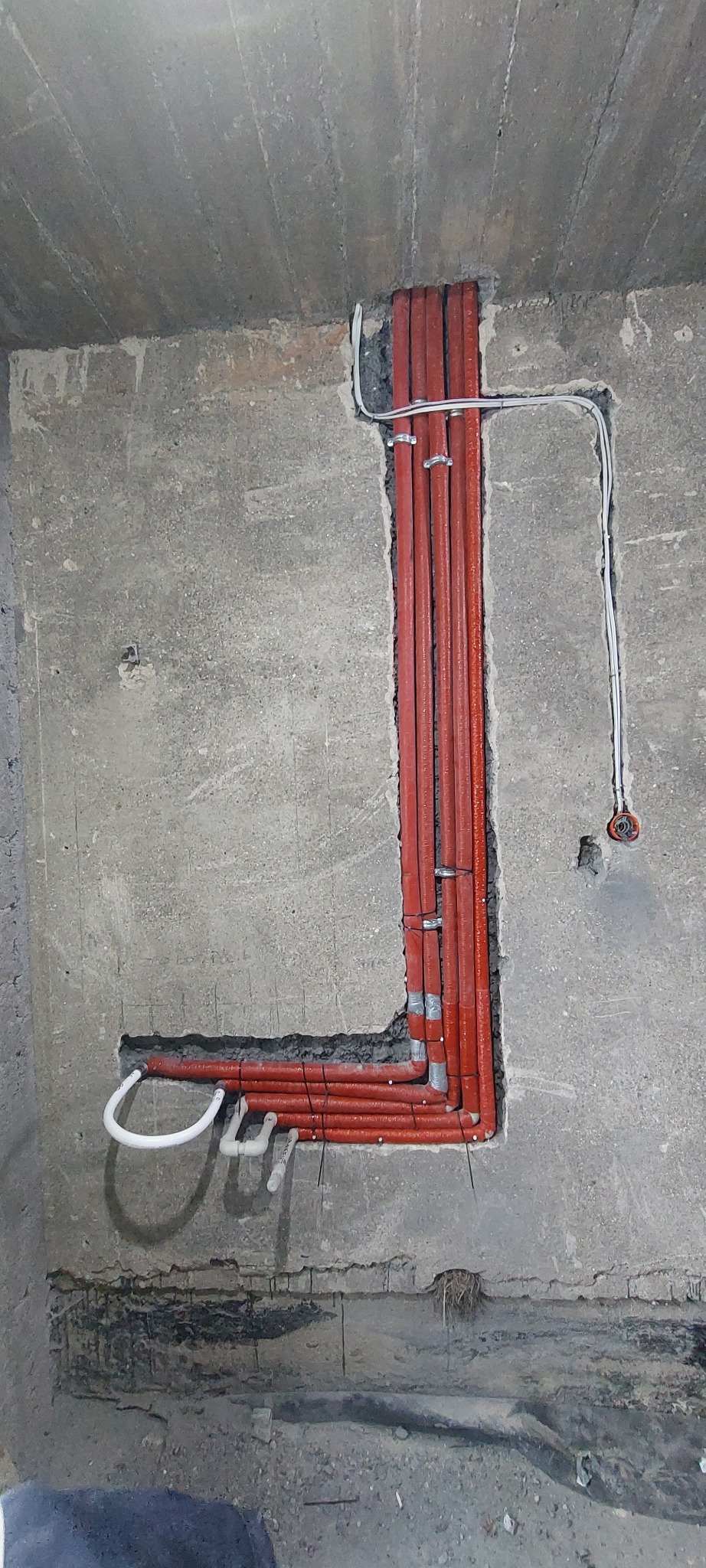 Czerwone rury ogrzewania podłogowego ułożone w bruzdach w surowym betonie, zabezpieczone metalowymi klamrami. Białe rurki i przewody elektryczne w tle.